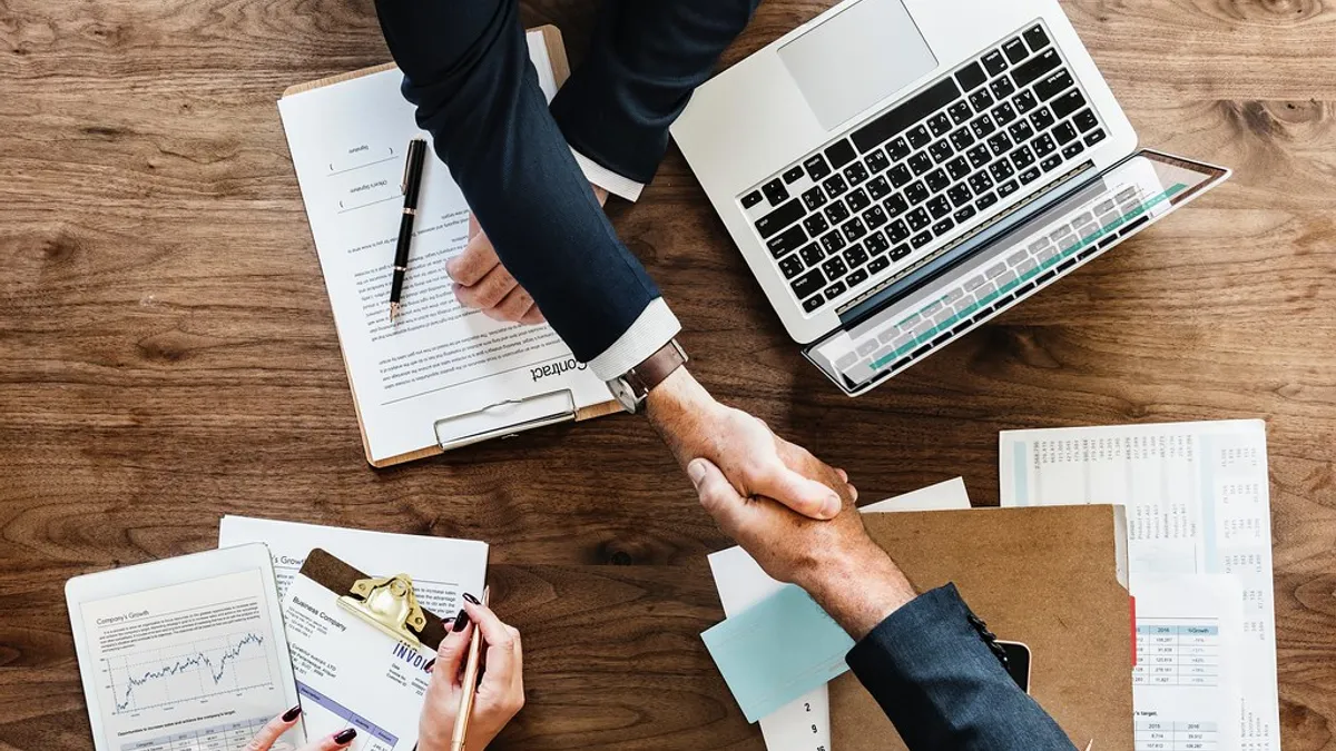 Twee mensen schudden handen boven een bureau met contracten en laptop